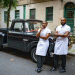A photo of two chefs leaning against a black truck outside their restaurant - photography competition submission