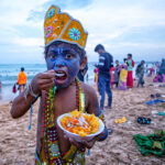 A photo of a young boy with face paint, wearing a colourful hat and multiple necklaces, eating a bowl of food on a busy beach - photography competition submission